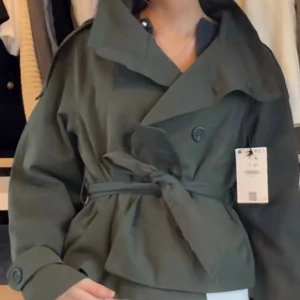 Kort trenchcoat med knytskärp - Trendig slutsåld grön trenchcoat från Bergslagen i storlek XS. Snygg kort trenchcoat med dubbelknäppning och brett knytskärp i midjan. Jackan har hög krage, axelklaffar och stora knappar. 