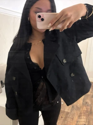 Mörkblå trenchcoat - Säljer en snygg svart trenchcoat med bred krage och stora dekorativa knappar framtill. Jackan har en boxig och kortare passform som ger en cool look. Perfekt att slänga över en outfit för att höja stilen. Materialet ser ut att vara bomull eller polyester.