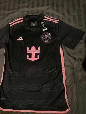 Inter Miami svart fotbollströja Adidas - Ej använd med tagg. Svart Inter Miami CF fotbollströja från Adidas med rosa detaljer och klubbmärke på bröstet. Tröjan har korta ärmar, rosa ränder på axlarna och rosa logga på magen. Tillverkad i lätt och ventilerande material, perfekt för träning eller match.