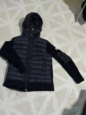 Svart pufferjacka från Moncler - Snygg svart pufferjacka från Moncler med vadderad front och huva. Jackan har stickade ärmar med Moncler-logga på vänster arm och dragkedjeficka. Perfekt för kalla dagar och ger en sportig vibe. Materialmix av dun och stickat tyg.