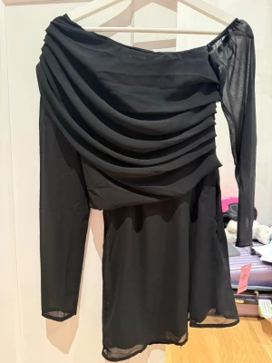 Svart oneshoulder kortklänning - Svart kortklänning med draperad front och transparent lång ärm på ena sidan. Klänningen har en asymmetrisk oneshoulder-design och är tillverkad i ett lätt, transparent syntetmaterial som ger en elegant look. Originalpris är ca 360kr