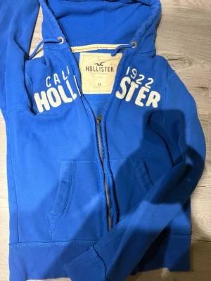 Sällsynt blå zip up hoodie från holister  - Sällsynt blå hoodie från Hollister med dragkedja och broderad vit text på bröstet. Modellen har huva med dragsko och två fickor framtill. Perfekt för en avslappnad och cool stil. Priset kan förhandlas 