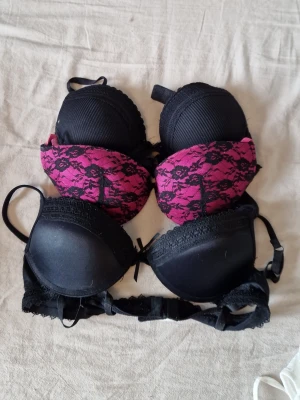 Tre bhar i spets, svart och rosa - Säljer tre bhar, två i helsvart med spetsdetaljer och en i rosa med svart spetsmönster. Alla har vadderade kupor och justerbara axelband. Perfekta för dig som gillar snygga och bekväma underkläder med lite extra detaljer.   Översta är i stl 75A  mitten i stl 75B sista i stl 75B.   50kr st. Alla är även push up.
