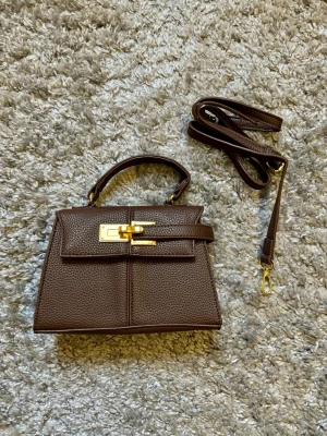 Mörkbrun väska - En stilren mörkbrun handväska med strukturerat läder och guldfärgade detaljer. Väskan har ett topphandtag och en avtagbar axelrem, samt ett elegant spänne framtill. Perfekt för dig som gillar klassiska och trendiga accessoarer.