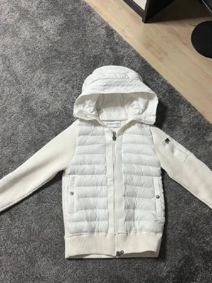 Vit cardiganjacka från Moncler - Snygg vit cardiganjacka från Moncler med vadderad framsida och huva. Ärmar och rygg är stickade i mjukt material, medan framsidan är quiltad. Jackan har dragkedja, fickor med tryckknappar och Moncler-logga på ärmen.
