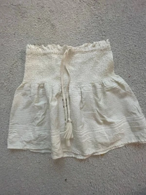 Vit kortkjol med knyt och volang - Supersöt vit kortkjol med smock i midjan och knytband med tofsar framtill. Kjolen har volangkant och ett lätt, luftigt tyg som passar perfekt till varma dagar. Enkel att matcha med olika toppar för en fräsch look. 