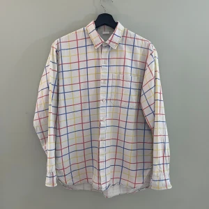 Vit rutig skjorta - Säljer en vit skjorta med färgglatt rutmönster i rött, blått och gult. Skjortan har långa ärmar och knappar framtill. Perfekt för en avslappnad stil eller en färgglad touch till vardagsoutfiten. Passar bra till både jeans och chinos!