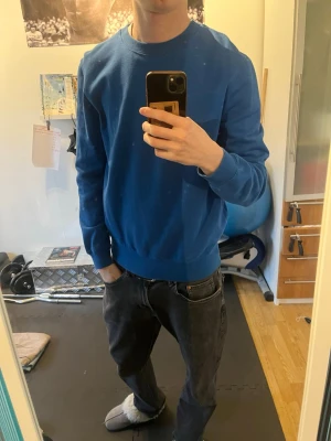 Blå sweatshirt - En stilren blå crewneck sweatshirt med rund hals och långa ärmar. Tröjan har ribbade muddar vid ärmslut och nederkant, perfekt för en avslappnad och enkel look. Passar bra till jeans eller joggers.