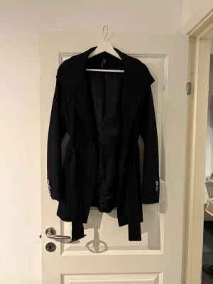 Svart kappa med skärp och stora slag - Stilren svart kappa med breda slag och knytskärp i midjan. Kappan har långa ärmar med dekorativa knappar vid ärmsluten och en klassisk, något figursydd passform. Perfekt för dig som vill ha en enkel men snygg look till hösten eller vintern. Priset är inte hugget i sten vid snabb affär!