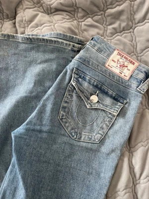 True Religion ljusblå bootcut jeans - Säljer ett par ljusblå jeans från True Religion, modell BECCA Mid Rise Bootcut. Jeansen har klassiska kontrastsömmar, ficklock med knapp bak och ikonisk logga på bakfickan. Tillverkade i mjukt denimtyg för en bekväm passform. Byxorna är använda fåtal gånger (2-3 gånger) då dom är i bra skick.