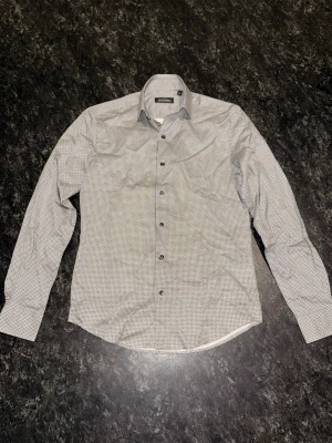 Vit mönstrad skjorta från Dressmann - Snygg vit skjorta från Dressmann med svart geometriskt mönster. Skjortan är som ny och oanvänd. Inga hål, fläckar eller skador. Den kommer att vara struken och tvättad i fint skick när du får den. Priset kan diskuteras.