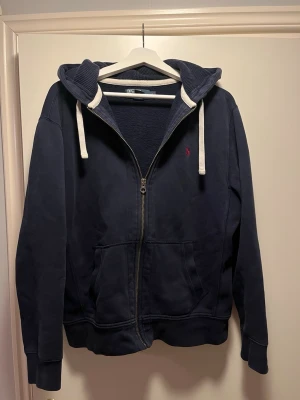 Ralph Lauren Zip Hoodie - Mörkblå hoodie med dragkedja från Polo Ralph Lauren. Klassisk design med vit dragsko i huvan, två fickor framtill och broderad röd logga på bröstet. Mjuk bomullskänsla och ribbade muddar vid ärmslut och nederkant. Bara att komma med frågor eller funderingar!