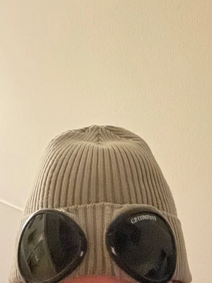 Grå C.P. Company goggle-mössa - Säljer en grå ribbstickad mössa från C.P. Company med ikoniska svarta goggle-linser framtill. Mössan har uppvikt kant och är tillverkad i mjukt material som sitter skönt på huvudet. Perfekt för dig som vill sticka ut med streetwear-stil.