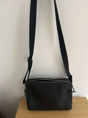 Svart axelväska från Lacoste - Snygg svart axelväska från Lacoste i rektangulär form med justerbar axelrem. Väskan har diskret Lacoste-logga framtill och är gjord i ett tåligt textilmaterial med struktur. Tvåvägsdragkedja i silverfärg ger en clean och modern look.