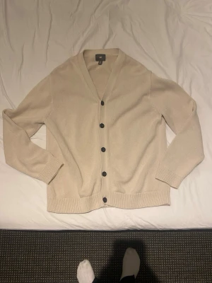 Beige kofta med svarta knappar - Stilren beige kofta med svarta knappar och v-ringad krage. Koftan har ribbade muddar vid ärmslut och nederkant, och är tillverkad i ett mjukt stickat material. Perfekt för lager-på-lager och enkel att matcha med olika outfits.