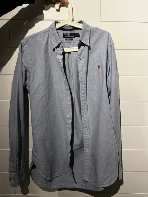 Polo Ralph Lauren skjorta - Klassisk ljusblå skjorta från Polo Ralph Lauren med slim fit-passform. Skjortan har knappar framtill, lång ärm och den ikoniska broderade loggan på bröstet. Tillverkad i mjuk bomull som känns skön mot huden. Perfekt för en clean och stilren look. Liten defekt på armbågen