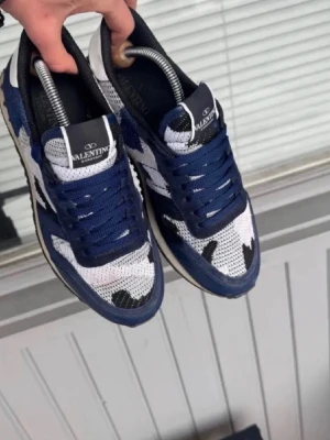 Valentino blå och vita sneakers - Snygga sneakers från Valentino i blått, vitt och svart med coolt camo-mönster. Ovandelen är i mesh och mocka, med vita detaljer och svarta inslag. Skorna har klassisk låg profil och vita sulor med blå detaljer. Perfekta för dig som vill sticka ut.