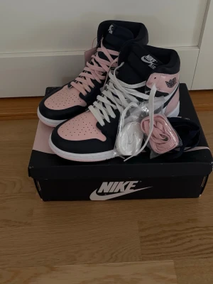 Nike Air Jordan 1 rosa/marinblå sneakers - Snygga Nike Air Jordan 1 sneakers i rosa och marinblå med vita detaljer. Skorna har högt skaft och kommer med extra skosnören i vitt, rosa och mörkblå. Klassisk vinge-logga på sidan och Nike Air-märke på plösen. Tillverkade i läder för en clean och stilren look.