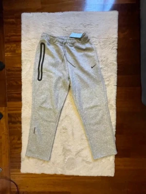 Grå Nike mjukisbyxor med dragkedja - Snygga grå mjukisbyxor från Nike med svart logga och en cool dragkedjedetalj längs vänster ben. Byxorna har resår i midjan och är tillverkade i mjukt, bekvämt material. Perfekta för chill eller träning.