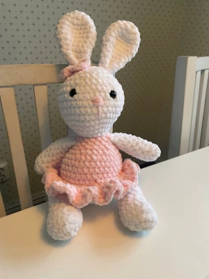 Virkad kanin i rosa klänning - Söt handvirkad vit kanin i mjukt garn med ljusrosa klänning och rosett. Kaninen har stora öron, svarta ögon och bär en rosa klänning med volangkant samt en liten rosett på ena örat. Perfekt som dekoration eller gosedjur. Virkad med kärlek