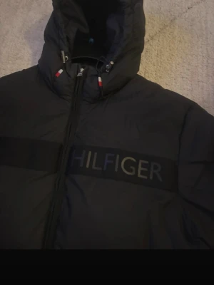 Svart pufferjacka från Tommy Hilfiger - Snygg svart pufferjacka från Tommy Hilfiger med stor logga över bröstet. Jackan har huva och detaljer i rött och vitt vid dragkedjan. Perfekt för kalla dagar och har en klassisk, puffig siluett. Materialet är slitstarkt och håller värmen bra.
