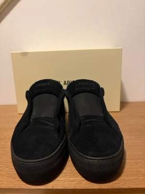 Svarta slip-on sneakers Axel Arigato - Säljer ett par helsvarta slip-on sneakers från Axel Arigato. Skorna är i mocka med diskret logga på sidan och har en chunky svart sula. Perfekta för dig som gillar minimalistisk och clean stil. Inga snören, bara att dra på sig och köra.