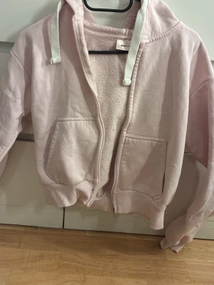 Ljusrosa hoodie med dragkedja - Mysig ljusrosa hoodie med dragkedja och två stora fickor framtill. Insidan är fodrad med teddy för extra värme. Huva med vita snören och ribbade muddar vid ärmslut och nederkant. Perfekt för en chill och avslappnad stil.