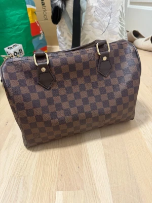 Handväska  - Snygg handväska i klassiskt Damier-mönster. Väskan är brun med rutigt tryck, har två handtag i mörkbrunt läder och guldfärgade metalldetaljer. Perfekt storlek för det viktigaste och stängs med dragkedja. Ikonisk och tidlös design. Bra skick 