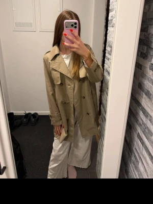 Beige trenchcoat med dubbelknäppning - Säljer en klassisk beige trenchcoat med bred krage och dubbelknäppning. Jackan har långa ärmar och snygga detaljer som axelklaffar och knappar framtill. Perfekt för lager-på-lager och stilrena outfits.