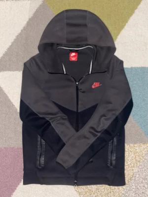 Svart Nike hoodie med dragkedja - Snygg svart hoodie från Nike med dragkedja framtill och två fickor med dragkedja. Klassisk Nike-logga i rött på bröstet och huva med paneldetaljer. Tillverkad i mjukt och bekvämt material, perfekt för en sportig och avslappnad stil.