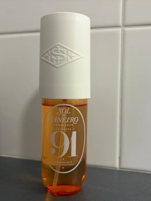 Sol de Janeiro Cheirosa 91 Parfymmist - Sol de Janeiro Cheirosa 91 är en parfymmist i en genomskinlig orange plastflaska med vit spray och lock. Flaskan har en modern, rundad form och tydlig logga framtill. Perfekt för dig som gillar fräscha och tropiska dofter.