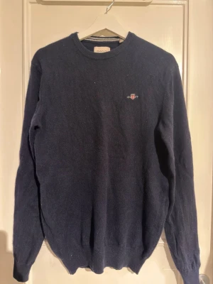 Mörkblå stickad tröja från Gant - Klassisk mörkblå stickad tröja från Gant med rund halsringning och diskret broderad logga på bröstet. Tröjan har långa ärmar och ribbade muddar vid ärmslut och nederkant. Perfekt till lager-på-lager och enkel att matcha.