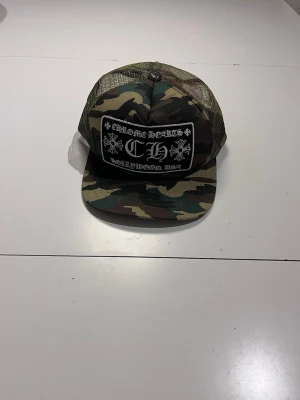 Camo keps från Chrome Hearts - Snygg keps i camomönster från Chrome Hearts med svartvit patch framtill och mesh på sidorna. Justerbar baktill och detaljer med Chrome Hearts-logga på insidan. Perfekt för dig som gillar streetwear och vill sticka ut.