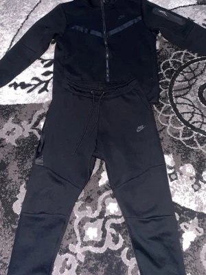 Svart Nike Tech Fleece hoodie - Snygg svart Nike Tech Fleece hoodie och byxor med dragkedja och huva. Diskret Nike-logga på bröstet och coola detaljer i form av svarta band över bröstet och på ärmen. Perfekt för en sportig och stilren look. Mjukt och bekvämt material.