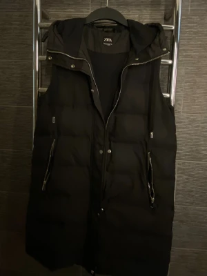 Svart lång dunväst från Zara - Säljer svart lång dunväst från Zara med huva och dragkedja framtill. Västen har två fickor med dragkedja. Perfekt till vår/sommar. 