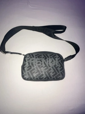 Svart axelväska från Fendi - Snygg svart axelväska från Fendi med justerbar rem och tydligt Fendi-logomönster över hela väskan. Väskan har en rektangulär form och är tillverkad i slitstarkt material med en cool streetstyle vibe. Perfekt för dig som vill ha en trendig och praktisk accessoar.