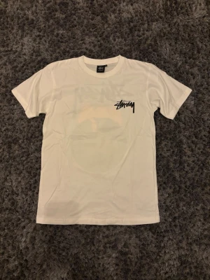 Vit Stüssy t-shirt med tryck - Vit t-shirt från Stüssy med svart logga på bröstet och stort grafiskt tryck på ryggen med en ängel och en biljardboll. Klassisk passform och rund hals. Perfekt för dig som gillar streetwear och vill sticka ut lite extra.
