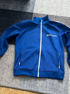 Blå zip hoodie från Palm Angels - Snygg blå hoodie från Palm Angels med vit dragkedja och vita detaljer längs ärmarna. Klassisk logga på bröstet och två fickor med dragkedja. Perfekt för en streetwear-look och enkel att matcha med jeans eller joggers.