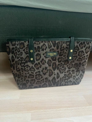 Leopardmönstrad shoppingväska Cahres & Heka - Snygg shoppingväska från Cahres & Heka med leopardmönster i brunt och svart. Väskan har svarta handtag med guldfärgade detaljer och är tillverkad i ett tåligt tygmaterial. Perfekt för dig som vill sticka ut med ett djurmönstrat statement.