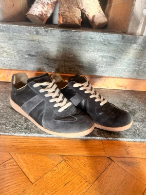 Mason margiela gats  - Säljer ett par snygga svarta sneakers med beige gummisula och vita snören. Skorna har en klassisk låg design och är tillverkade i mocka och läder. Perfekta för dig som gillar stilrena och tidlösa skor. Ena skon sönder därav priset. 