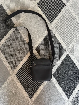 Svart axelväska från BOSS - Snygg svart axelväska från BOSS i stilren design. Tillverkad i slätt konstläder med justerbar axelrem och en praktisk ytterficka med dragkedja. Diskret BOSS-logga framtill och metall-detaljer för en clean look.