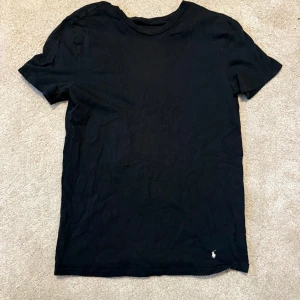 Svart t-shirt från Ralph Lauren - Klassisk svart t-shirt från Ralph Lauren med liten broderad logga i vitt nedtill. T-shirten har rund halsringning och korta ärmar. Perfekt basplagg med stilren design och skön passform.