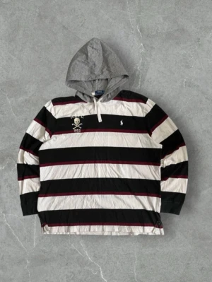 Randig hoodie från Polo Ralph Lauren - Snygg hoodie med breda svarta, vita och vinröda ränder från Polo Ralph Lauren. Grå huva med dragsko och broderad dödskalle med texten R.L.F.C. 1923 på bröstet. Klassisk Polo-logga på ena sidan. Perfekt för en avslappnad streetwear-look.