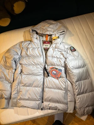 Parajumpers pharell sheen jacka - Snygg silverfärgad dunjacka från Parajumpers med huva, två sidofickor med dragkedja och klassisk patch på ärmen. Jackan har quiltad design och detaljer i orange och gul vid halsen. Perfekt för kalla vinterdagar.M passar S. Kvitto med följer inköpt för 6200kr då fick den som julgåva 