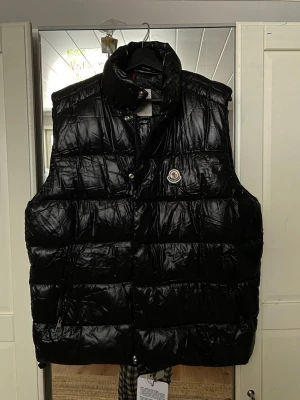 Svart dunväst från Moncler - Snygg svart dunväst från Moncler med hög krage och dragkedja framtill. Västen har två sidofickor med dragkedja och det klassiska Moncler-logotypen på bröstet. Tillverkad i glansigt material som ger en modern look.