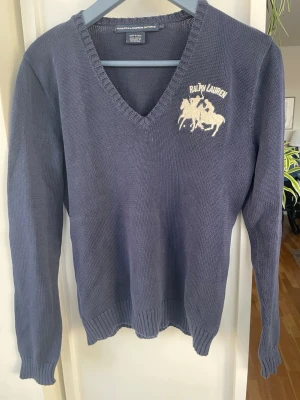 Blå v-ringad vintage tröja från Ralph Lauren, stel L - Mysig blå stickad tröja från Ralph Lauren med v-ringning och lång ärm. Tröjan har en vit broderad logga med hästmotiv på bröstet och ribbade muddar vid ärmslut och nederkant. Perfekt för en stilren och avslappnad look. Använd vid enstaka tillfällen och är i väldigt bra skick 
