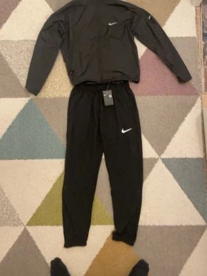 Svart träningsset från Nike - Snyggt svart träningsset från Nike med matchande jacka och byxor. Båda plaggen har vita Nike-loggor och är tillverkade i ett lätt, funktionellt material som passar perfekt för löpning eller gym. Jackan har dragkedja och byxorna har resår i midjan.