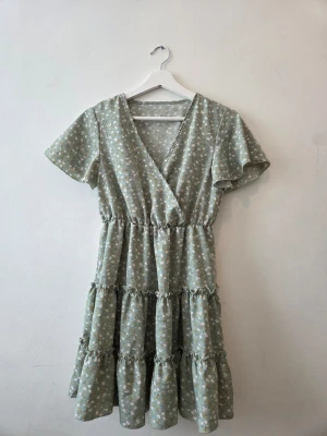 Grön blommig klänning - Söt grön kortklänning med blommönster. Klänningen har volangdetaljer, korta ärmar och resår i midjan för en bekväm passform. Perfekt för vår och sommar!
