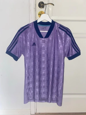 Lila Adidas träningströja - En lila tränings t-shirt från Adidas med snygga detaljer och klassiska ränder på axlarna. Tröjan har ett diskret rutmönster i tyget, V-ringad hals och korta ärmar. Perfekt för träning eller sportiga outfits. Storlek S, använd en gång