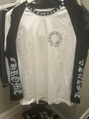 Chrome Hearts långärmad t-shirt - Säljer en svartvit långärmad t-shirt från Chrome Hearts med stora logotexter på ärmarna och ett stort tryck på ryggen. Klassisk baseballmodell med svart krage och ärmar, samt vit kropp. Perfekt för dig som gillar streetwear och vill sticka ut.